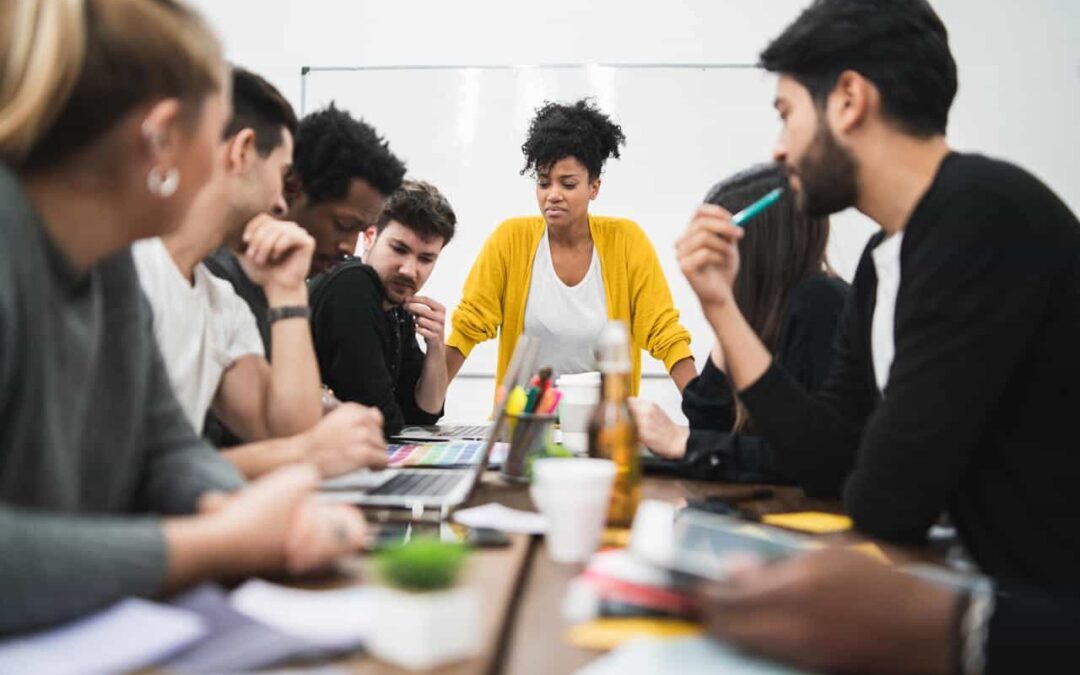 Imagem de uma mesa de reunião cm várias pessoas em volta e notebooks em cima da mesa.