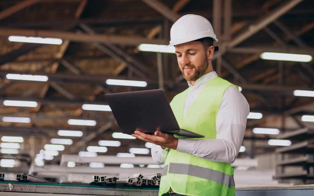 Homem com capacete de proteção e colete sinalizador, com um notebook aberto. A foto foi tirada em um galpão de distribuidora.